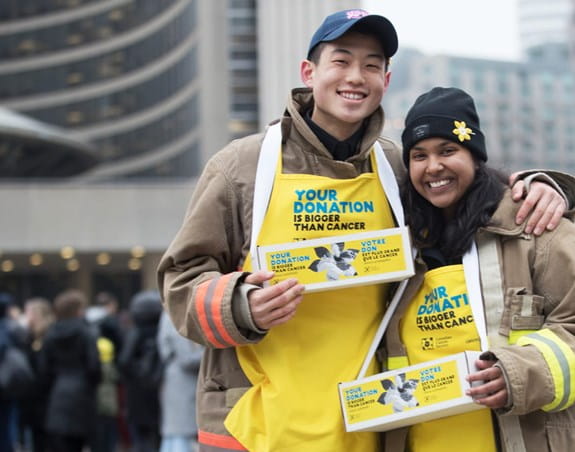 Young man and young woman wearing yellow Daffodil campaign aprons, holding daffodil pin boxes at City Hall, Toronto